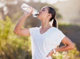 heureuse femme brune en t-shirt blanc bouteille d'eau potable à l'extérieur par une journée ensoleillée après l'entraînement