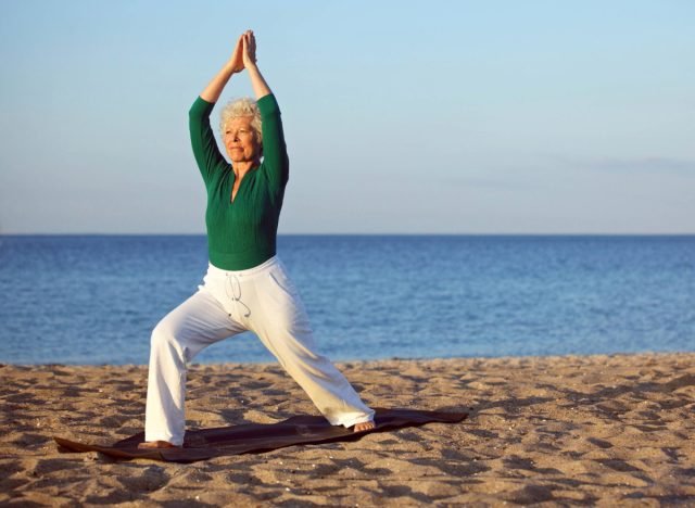 femme mûre en haut vert à manches longues et pantalon blanc faisant du yoga à l'heure d'or sur la plage