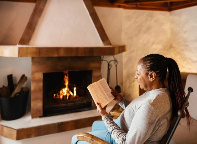 femme mûre assise sur une chaise lisant un livre près de la cheminée