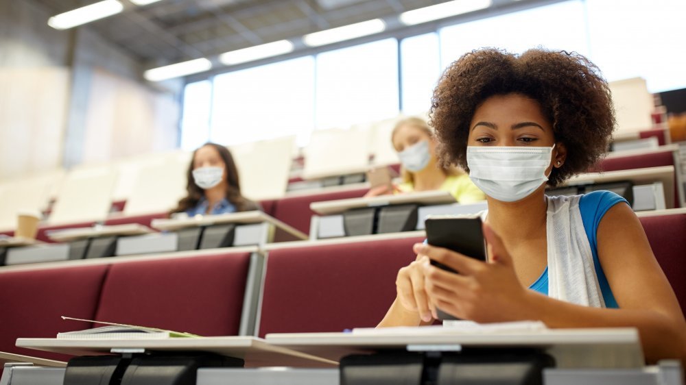 Une jeune femme dans une salle de classe d'université porte un masque facial et vérifie son téléphone