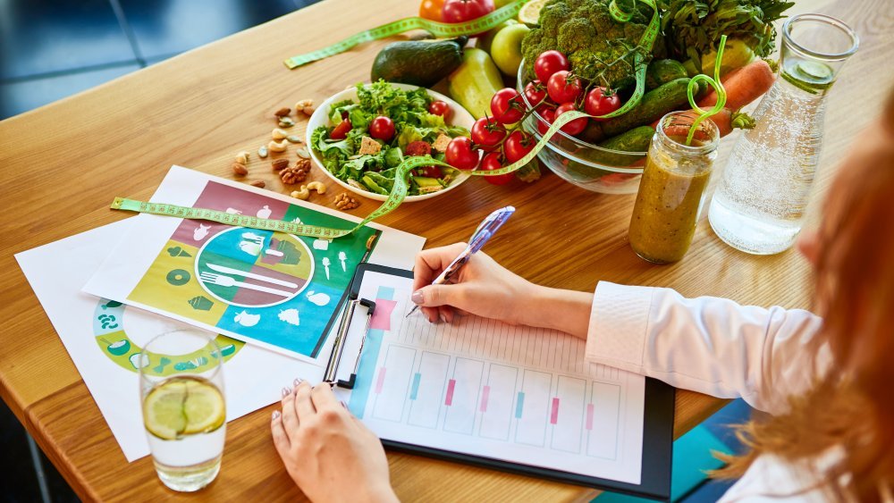 Une diététicienne en uniforme médical avec un ruban à mesurer travaille sur un régime alimentaire assis avec différents ingrédients alimentaires sains dans le bureau vert en arrière-plan. Concept de perte de poids et de bonne nutrition. Régime inversé. 