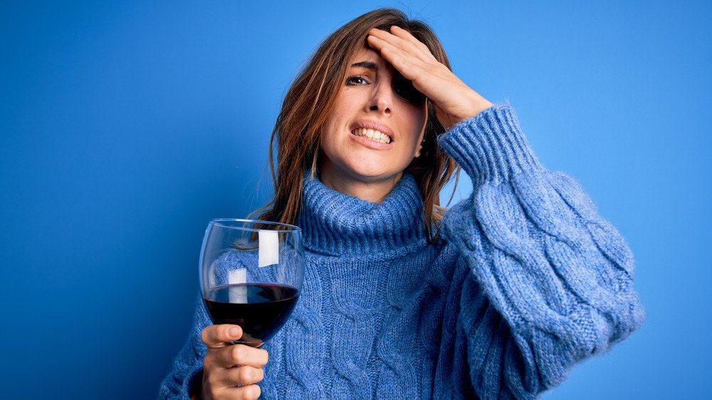 Personne en pull bleu grimaçant et tenant un verre de vin rouge avec la main sur la tête